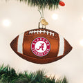 A brown football-shaped ornament with the Alabama Crimson Tide logo hangs on a Christmas tree, surrounded by green pine needles and blurred holiday lights in the background.