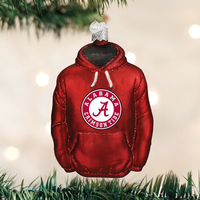A Christmas tree ornament shaped like a red hoodie with the Alabama Crimson Tide logo hanging from a branch, with blurred white lights in the background.