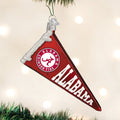 A red Alabama Crimson Tide pennant ornament with white lettering and logo hangs on a Christmas tree, surrounded by blurred white lights.