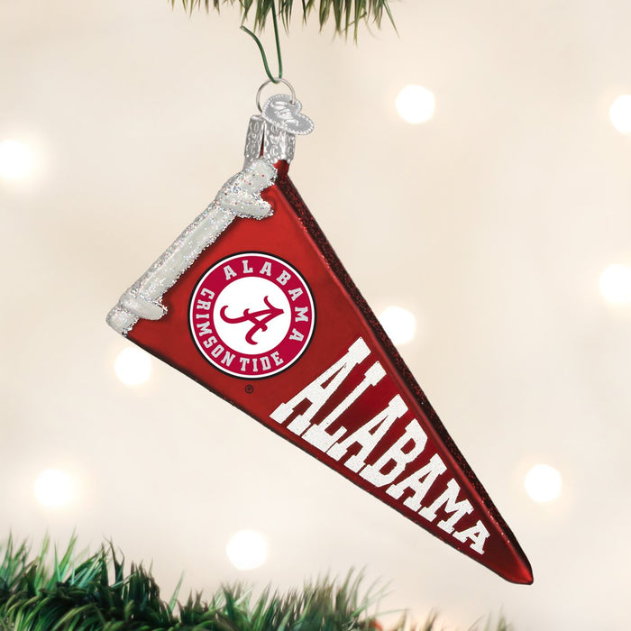 A red Alabama Crimson Tide pennant ornament with white lettering and logo hangs on a Christmas tree, surrounded by blurred white lights.