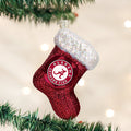 A glittery red Christmas stocking ornament with a white top hangs on a tree branch. The stocking features the Alabama Crimson Tide logo in the center. The background shows blurred white lights.