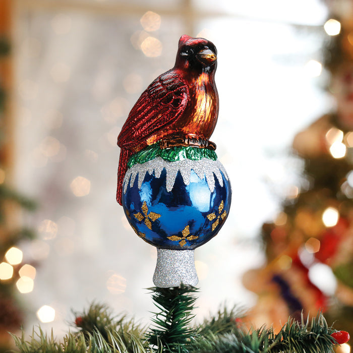 A red cardinal ornament sits atop a blue and silver globe with gold accents, used as a Christmas tree topper. Blurred holiday lights and decorations are in the background.