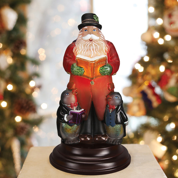 A festive figurine of Santa Claus in a red coat holding a book titled Carols, accompanied by two penguins dressed in scarves, also holding books, standing on a round base with blurred holiday lights in the background.