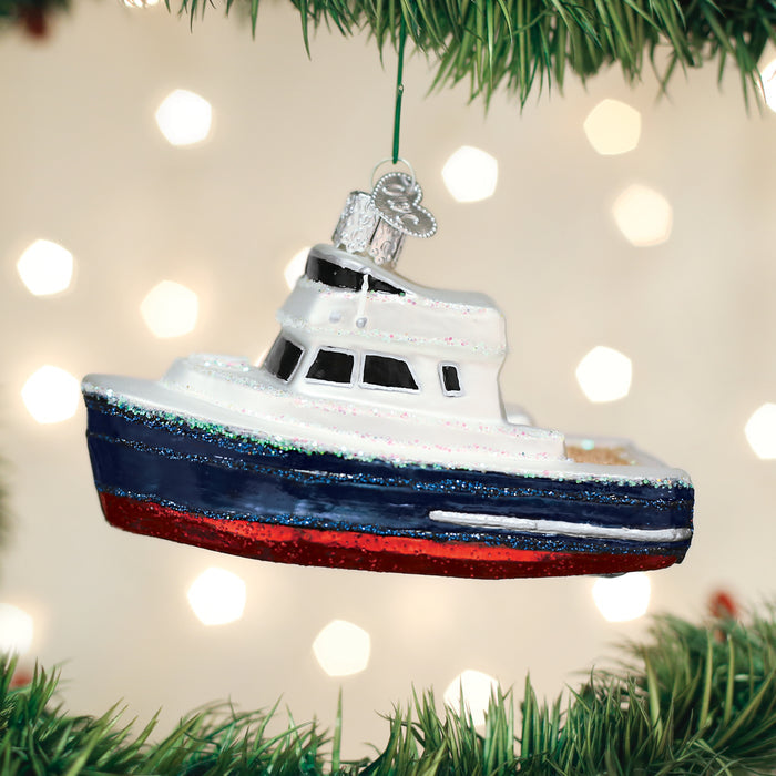 A Christmas tree ornament shaped like a small boat with white, blue, and red colors hangs among green tree branches, with blurred white lights in the background.