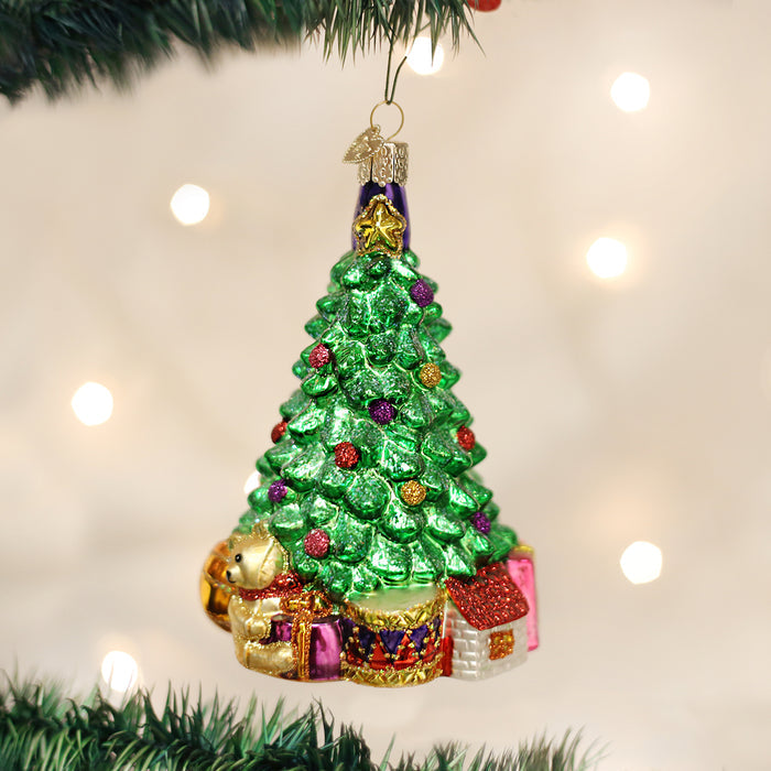 A shiny Christmas tree ornament decorated with colorful baubles, small houses, and a gold dog sits against a blurred background of white holiday lights and green tree branches.