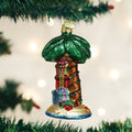 A colorful glass ornament shaped like a palm tree with presents at its base hangs from a green Christmas tree branch, with soft white lights blurred in the background.