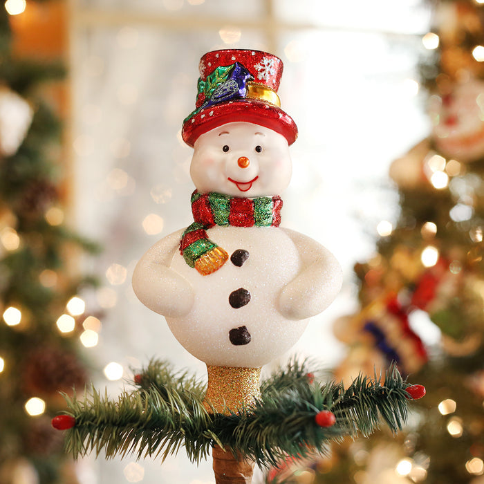 A cheerful snowman tree topper with a red glittery hat, striped scarf, and buttons stands on top of a Christmas tree, surrounded by blurred holiday lights and decorations.