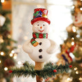 A cheerful snowman tree topper with a red glittery hat, striped scarf, and buttons stands on top of a Christmas tree, surrounded by blurred holiday lights and decorations.