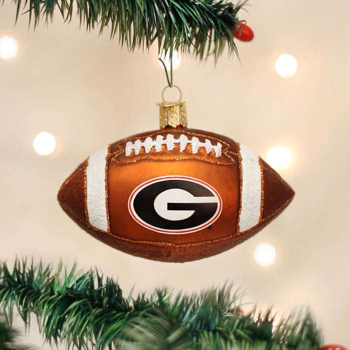 A football-shaped Christmas ornament with the University of Georgia G logo hangs from a green Christmas tree branch, with soft white lights in the background.