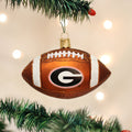 A football-shaped Christmas ornament with the University of Georgia G logo hangs from a green Christmas tree branch, with soft white lights in the background.