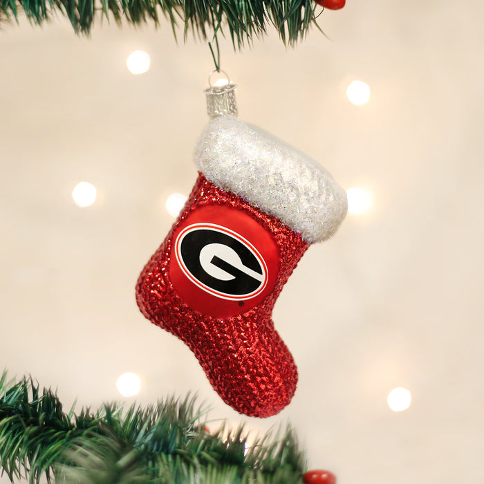 A red Christmas stocking ornament with a white glittery cuff and the Georgia Bulldogs G logo hangs from a green Christmas tree branch, with soft white lights in the background.