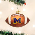 A football-shaped Christmas ornament with the University of Michigan M logo hangs from a green tree branch with a blurred, light background.