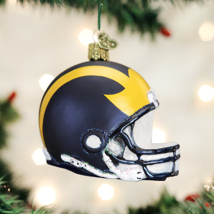 A Christmas tree ornament shaped like a football helmet with a navy blue and yellow design, hanging on a tree with blurred lights in the background.