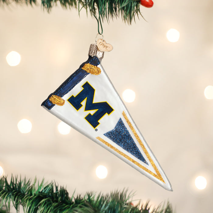 A white pennant-shaped ornament with a blue and yellow M hangs on a Christmas tree, decorated with gold and blue glitter accents. Blurred lights glow in the background.