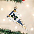 A white pennant-shaped ornament with a blue and yellow M hangs on a Christmas tree, decorated with gold and blue glitter accents. Blurred lights glow in the background.