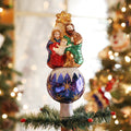 A Christmas tree topper featuring a glittery ornament with a figurine of the Holy Familyâ€”Mary, Joseph, and baby Jesusâ€”sitting on top, surrounded by Christmas tree branches and festive decorations.