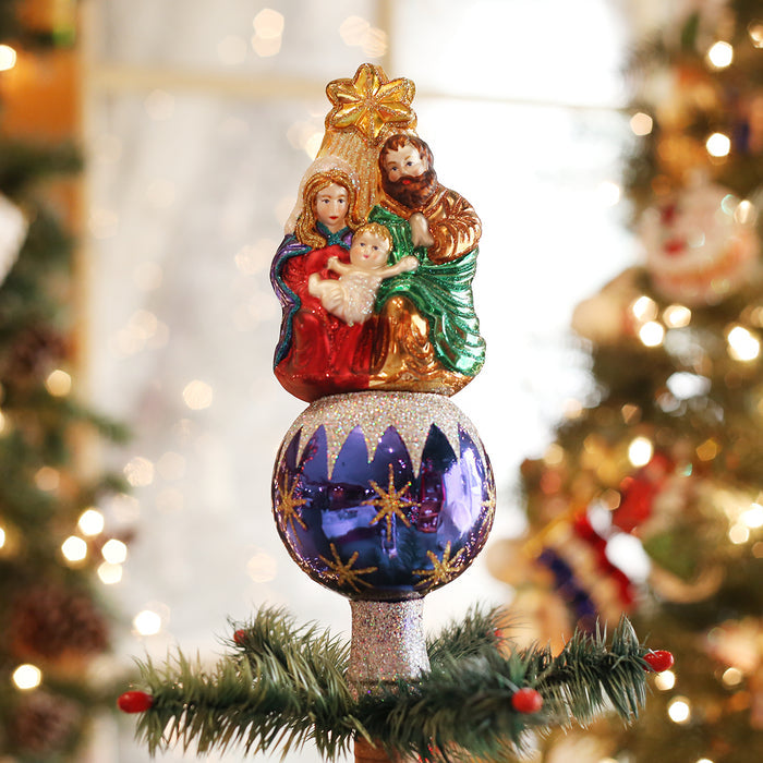 A Christmas tree topper featuring a glittery ornament with a figurine of the Holy Familyâ€”Mary, Joseph, and baby Jesusâ€”sitting on top, surrounded by Christmas tree branches and festive decorations.