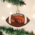 A football-shaped Christmas ornament with the Oregon State Beavers logo hangs on a green tree branch with blurred lights in the background.
