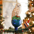 A white owl ornament with gold accents sits atop a blue glittery sphere and green leaves, perched on the top of a Christmas tree with blurred holiday lights and decorations in the background.
