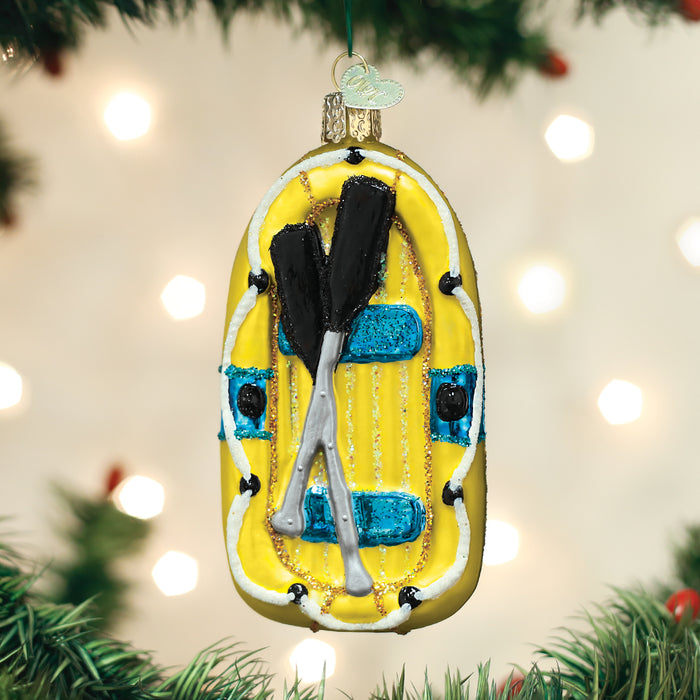 A yellow inflatable raft ornament with two black and silver paddles crossed on top hangs on a Christmas tree, with blurred white lights in the background.