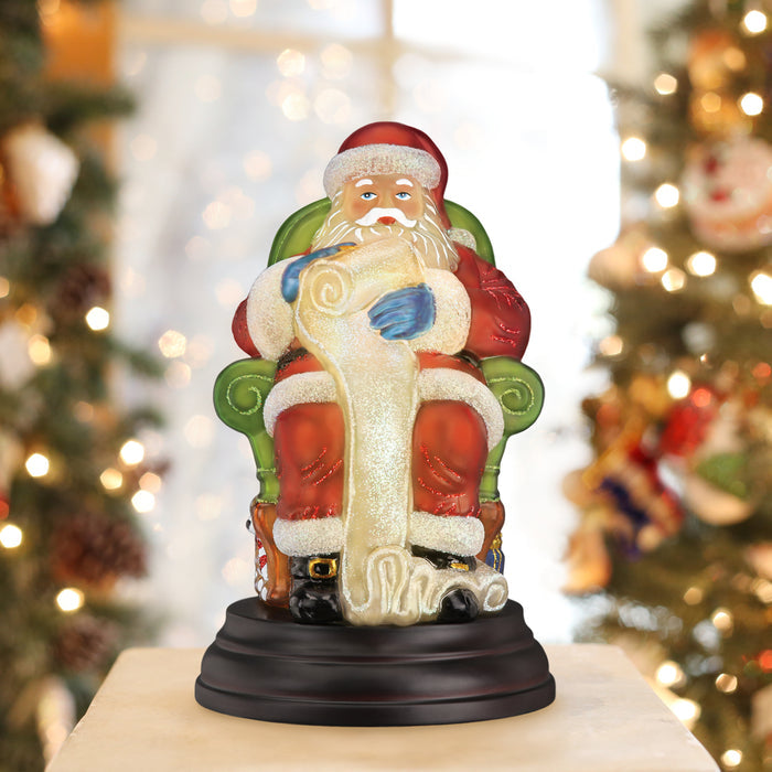 A festive figurine of Santa Claus sitting on a green chair and holding a long scroll is displayed on a table, with blurred Christmas trees and lights in the background.