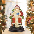 A festive figurine of Santa Claus sitting on a green chair and holding a long scroll is displayed on a table, with blurred Christmas trees and lights in the background.