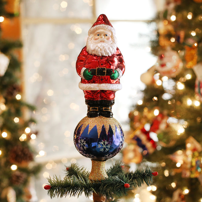 A glass Christmas tree topper shaped like Santa Claus stands on a glittery blue and gold ornament ball, with decorated Christmas trees and twinkling lights softly blurred in the background.