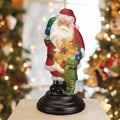 A festive Santa Claus figurine holding a teddy bear and standing beside a smiling child in green, displayed on a black base with decorated Christmas trees and warm lights in the blurred background.