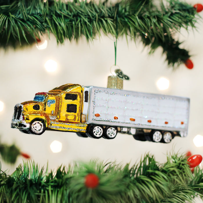 A glittery yellow and silver semi-truck ornament hangs from a green Christmas tree branch, with blurred white lights in the background.