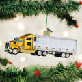 A glittery yellow and silver semi-truck ornament hangs from a green Christmas tree branch, with blurred white lights in the background.