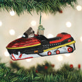 A red, black, and silver snowmobile ornament hangs from a Christmas tree branch, with blurred white lights in the background.