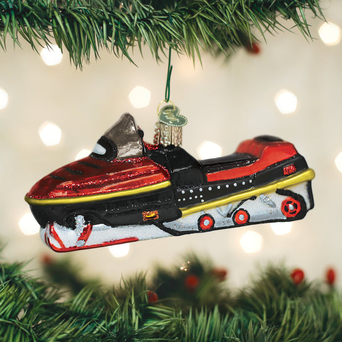 A red, black, and silver snowmobile ornament hangs from a Christmas tree branch, with blurred white lights in the background.