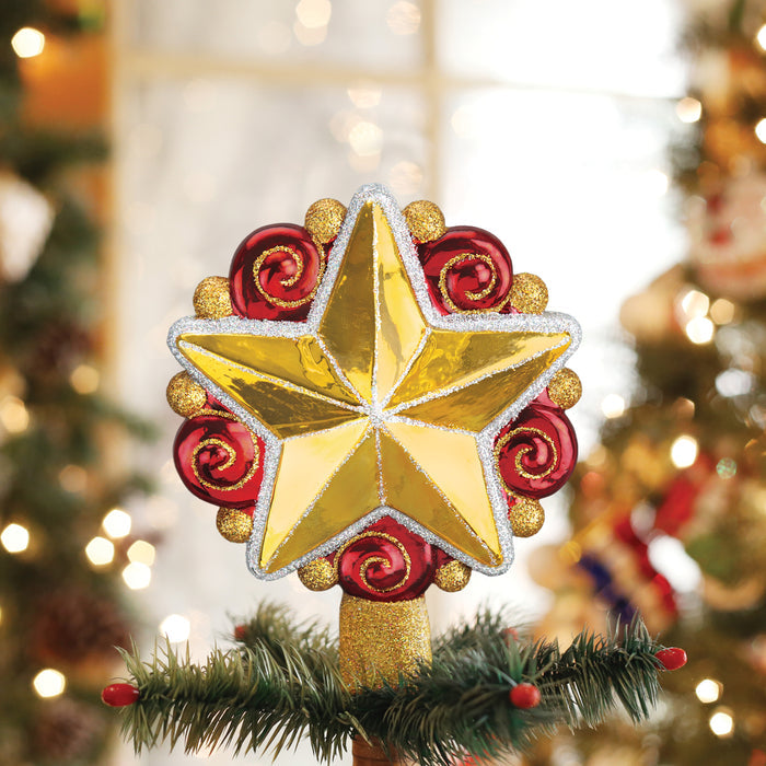 A shiny gold star tree topper with red spiral accents sits on top of a Christmas tree branch, with blurred festive decorations and lights in the background.