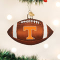 A Christmas tree ornament shaped like a football with a large orange letter T on it, hanging from a branch with blurred lights in the background.