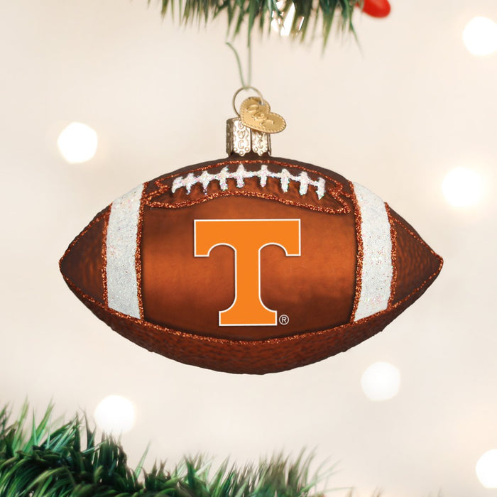 A Christmas tree ornament shaped like a football with a large orange letter T on it, hanging from a branch with blurred lights in the background.