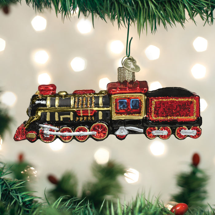 A shiny, colorful train ornament with red, black, and gold details hangs from a Christmas tree branch, with blurred holiday lights in the background.
