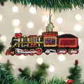 A shiny, colorful train ornament with red, black, and gold details hangs from a Christmas tree branch, with blurred holiday lights in the background.