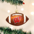 A Christmas tree ornament shaped like a football with a red W logo, hanging on a green tree branch with blurred white lights in the background.