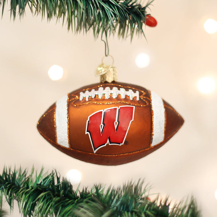 A Christmas tree ornament shaped like a football with a red W logo, hanging on a green tree branch with blurred white lights in the background.